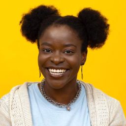 A smiling Black woman with two afro puffs sits cross legged on a floor wearing khaki shorts, a tan cardigan, and a shirt that reads "Louisiana girls are sunshine mixed with a little bit of hurricane."