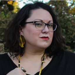 Jen, a light-complected femme presenting person with black glasses, brown, wavy, shoulder-length hair, sits in a park with stone masonry and trees behind hir. Jen’s left fist is raised in the air, exposing tattoos and a Native designed turquoise, yellow, red, and white bangle bracelet. Hir right hand is holding onto one side of a Native made yellow, black, and white beaded medallion with the Crushing Colonialism logo in the center. Jen is wearing a black, short sleeve shirt and large, yellow earrings.