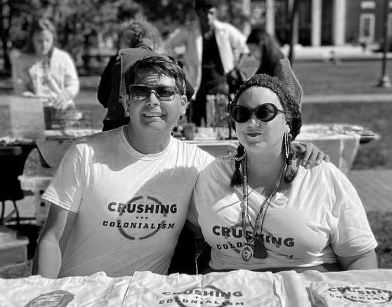 A black and white photo of two Native people wearing sunglasses and Crushing Colonialism tshirts sit beind a table of Crushing Colonialism merchandise. They’re outside and people are in the background.