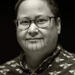 A picture of a smiling Native person in glasses with face tattoos and a button-up collared shirt is on a yellow background.
