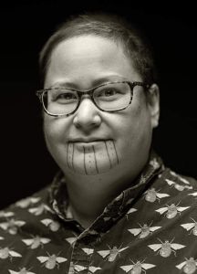 A picture of a smiling Native person in glasses with face tattoos and a button-up collared shirt is on a yellow background.
