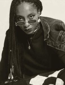 A Halpulaar / Lokono person wearing black sunglasses kneeling wearing black and white Fulani chayah trousers, a black top, a black jean jacket & black boots looking up at the camera. Rivolta is wearing a cowrie shell embedded in silver jewelry set with long brown braids.