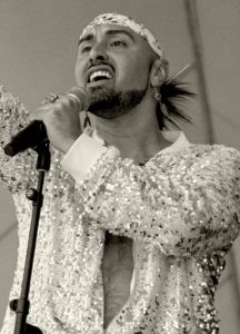 A picture of a singing Native person holding a mic in one hand with the other arm raised, wearing a loose silver and white long sleeve top open to the belly button and a matching headband.