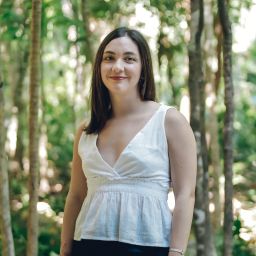 Isabel Riofrío is a Latino woman, standing in the middle of a garden full of trees. She is wearing a white top and blue trousers, and part of her cherry blossom tattoo is exposed. She is smiling as she looks at the camera.