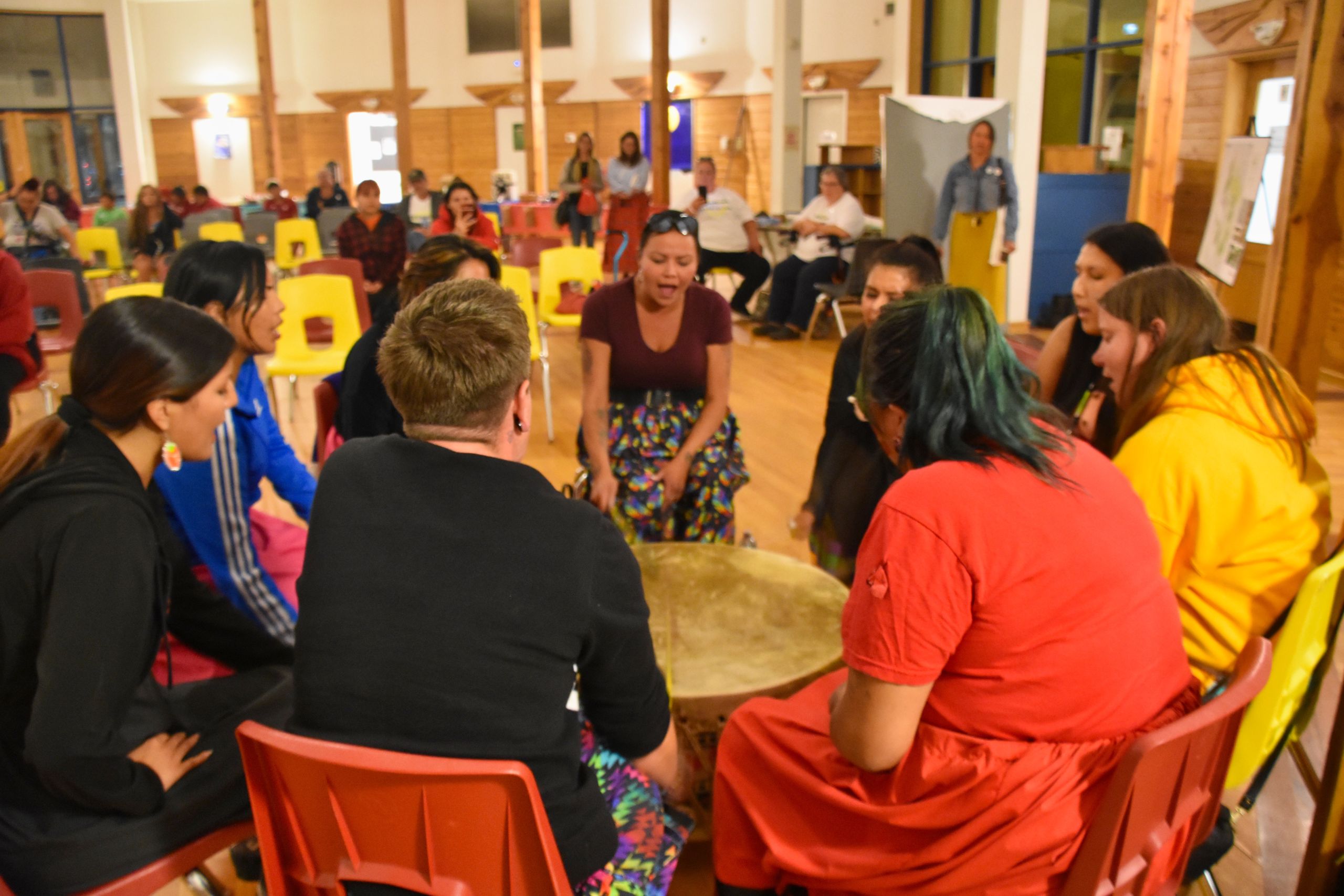 Photo of nine Indigenous women and Two-Spirits sitting around a large drum singing and hitting the drum with drumsticks. An audience watches in the background. Photo credit Jen Deerinwater.