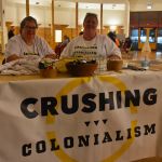 Photo of two people sitting at the welcoming table with a large Crushing Colonialism banner.