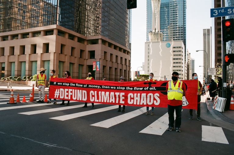A group of people blocks a major downtown “Los Angeles” intersection crosswalk while holding a long horizontal banner that reads, to protest climate disaster financiers. Photo credit: Steph Viera, November 25, 2022