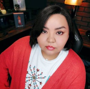 Johnnie Jae, a native women with short black hair and brown eyes staring straight ahead while wearing a coral lipgloss, a white graphic tee and a coral cardigan sweater.