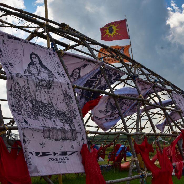 Photo of a longhouse frame covered with red dresses and banners, at Camp Morgan on Treaty 1 territory (“Winnipeg, Manitoba, Canada”) taken on August 8, 2023. Photo credit Jen Deerinwater