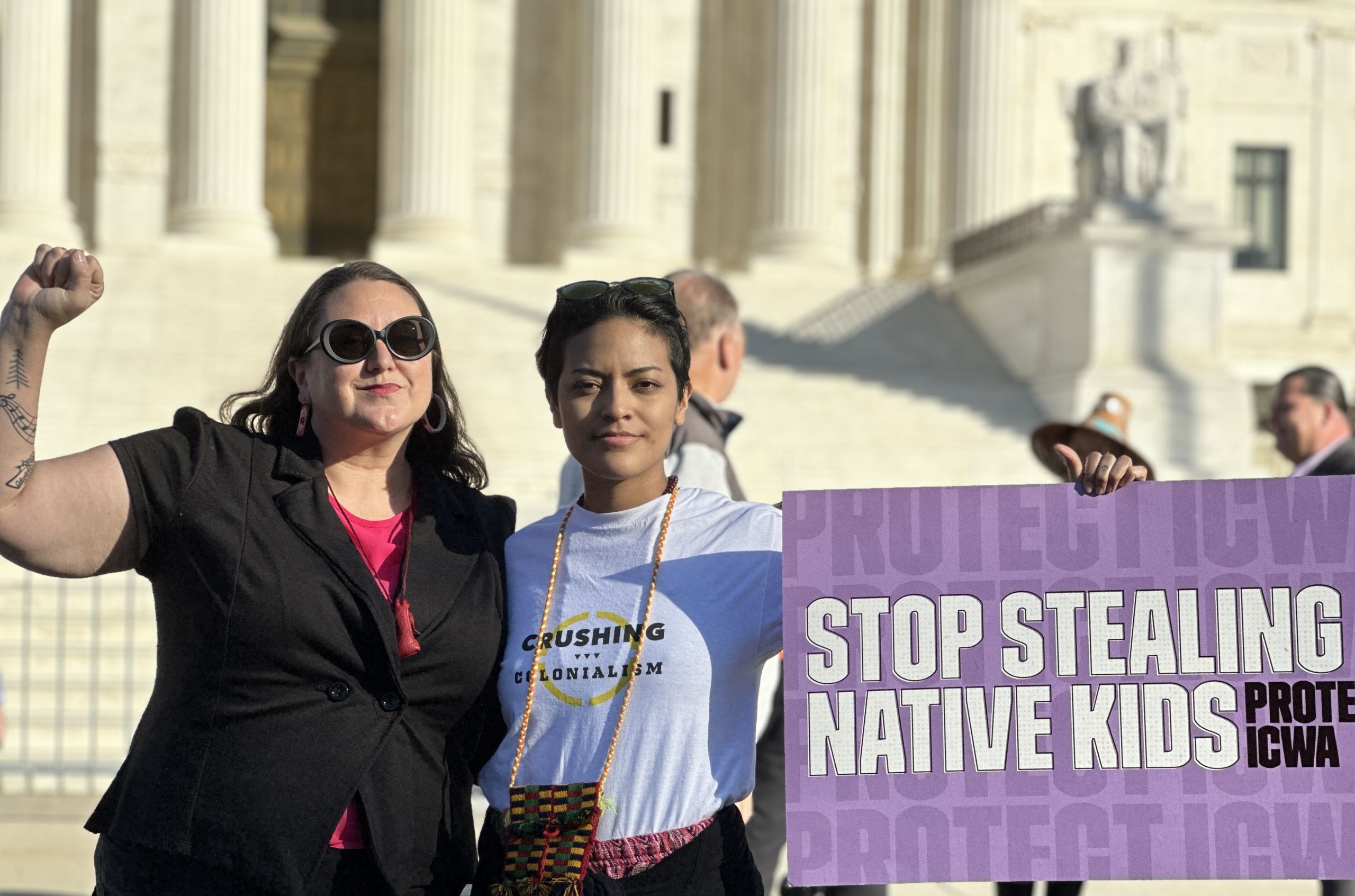 Two Indigenous people stand together in front of the US Supreme Court. One has their fist in the air while the other holds a sign that reads “Stop stealing Native kids.”