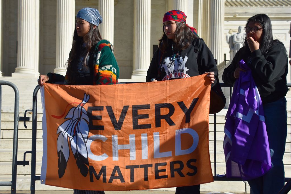 Three Native youth stand together in front of the US Supreme Court. They’re holding an orange flag with a feather on it that reads “Every Child Matters.”