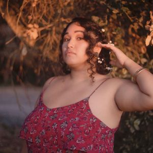 Photo of Solange Aguilar. Solange is standing at a three-quarter angle and is tucking their hair back behind their ear with their left hand. They have two braids with small white flowers in them and they're wearing a red maxi floral print spaghetti strap dress with gold bangles. They are looking into the camera with their head lifted slightly and are standing in front of an out-of-focus natural background. It is golden hour and the lighting is a light orange color with their face illuminated.