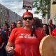 Photo of an Indigenous woman in a red t-shirt that reads "My sisters are warriors", She's holding sage and a drum. Behind her are other Indigenous women and allies.