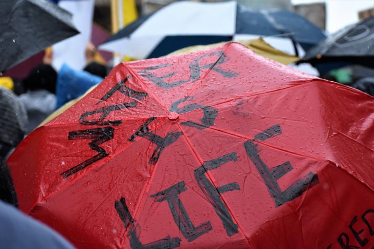 Close up photo of the top of a rain covered red umbrella that reads in black marker “Water is Life.” It is surrounded by numerous black umbrellas in the background.