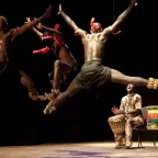 The dancers of the group Netos de Bandim are performing on stage. The photo shows two men on the background playing a musical instrument while three dancers jump to the beat. The photo is credited Grupo Cultural Netos de Bandim.