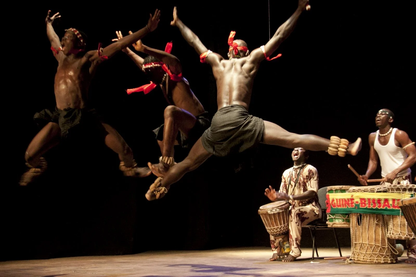 The dancers of the group Netos de Bandim are performing on stage. The photo shows two men on the background playing a musical instrument while three dancers jump to the beat. The photo is credited Grupo Cultural Netos de Bandim.