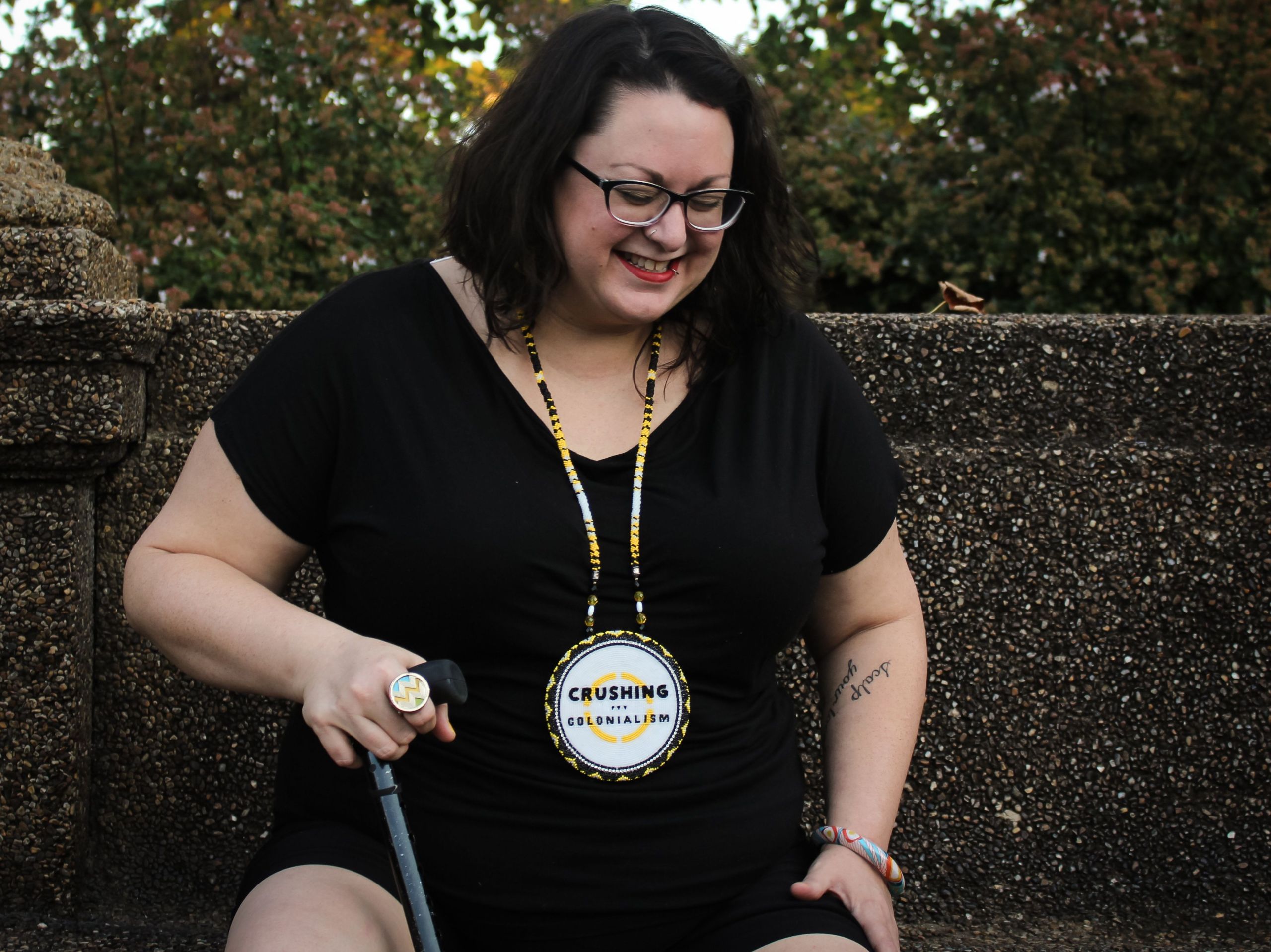 Jen is posing for a photo in Malcolm X Park on Piscataway lands ("Washington D.C, US"). In the photo Jen is sitting with her hand on her cane and is bent over laughing. It's a moment of joy caught during a photo shoot.. She is wearing a black t-shirt and shorts with a Native designed medallion of the Crushing Colonialism logo. Photo by Eleanor Goldfied.
