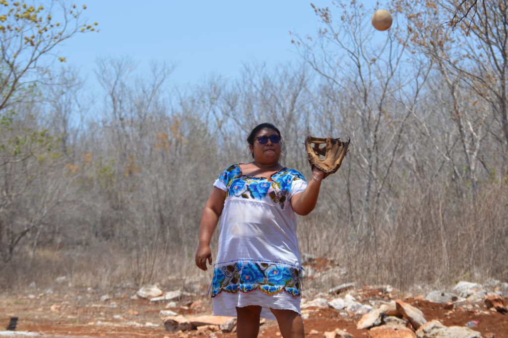 The photo shows Yessica Yazmin Diaz Canul warming up before the softball game against the Águilas de San Antonio, in Halachó, in México's Yucatán peninsula. She has a baseball glove on her right hand and the ball coming at her. She is wearing a traditional white and blue Huipil dress, and sunglasses to protect from the sun.