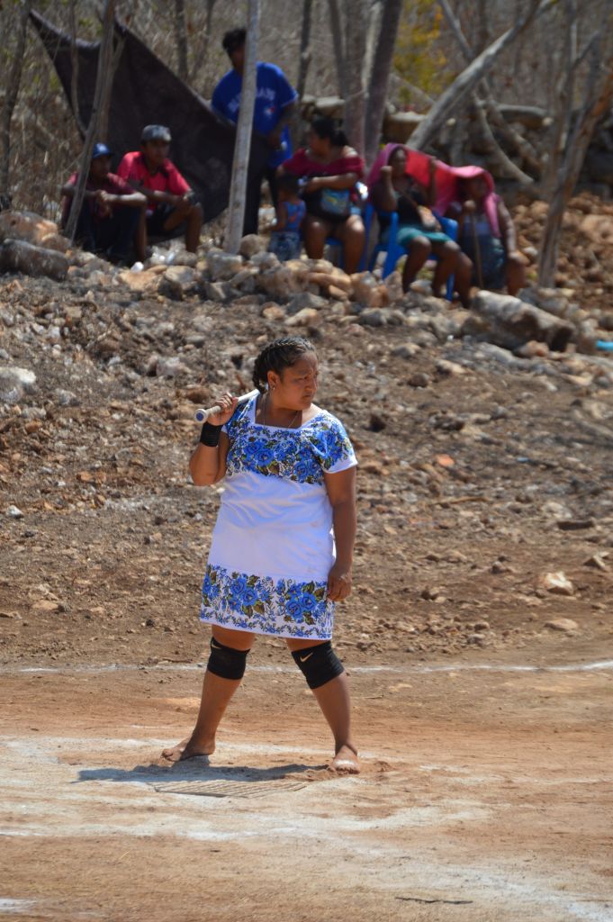 The photo shows Daniela Patrocinia settling into the batter's box. She has the bat on one hand, and is looking straight at the pitcher. She is barefoot and playing in the traditional Huipil dress, colored white and blue.
