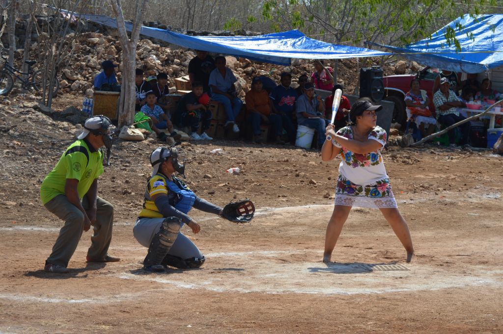 The photo shows Ceci Chan ready to swing during the game against the Águilas de San Antonio, in the municipality of Halachó, in México's Yucatán peninsula. Ceci is holding the bat with both hands and barefoot. Behind, people are watching the game underneath a tent to protect from the sun.