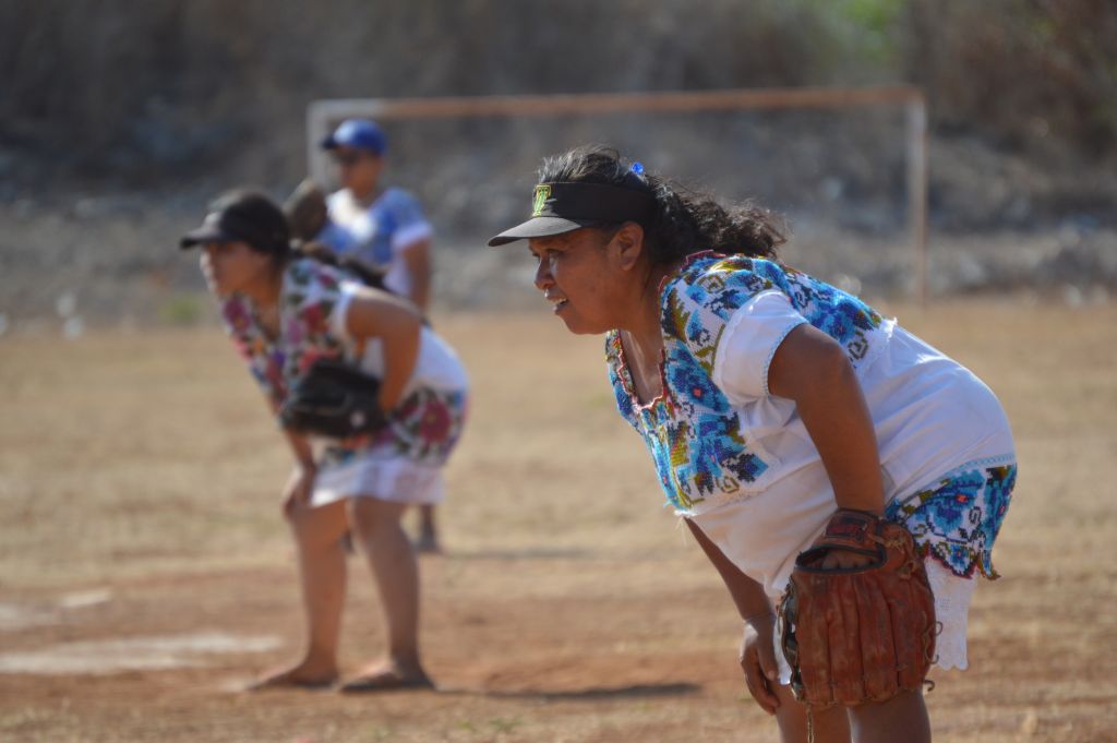 The photo shows María Enedina Canul guarding first base. In the background, her teammates are concentrated in the game.