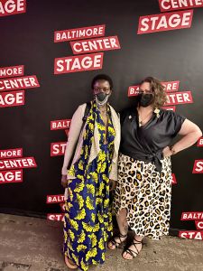 DeAnna Rhodes, wearing a long black and yellow dress with a light cardigan poses with Jen DeerinWater, wearing a Black V-neck blouse and leopard spotted skirt against a Black backdrop with the words Baltimore Center Stage