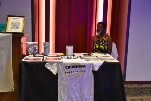 DeAnna Rhodes stands behind the Crushing Colonialism exhibit table that has a Crushing Colonialism tee shirt draped over a chair.