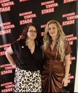 Eleanor Goldfield, a blonde woman wearing a brown dress, is posing next Jen Deerinwater, who is wearing a black v-neck blouse and leopard spotted skirt against a black backdrop that says Baltimore Center Stage.