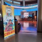 A photo of the entrance to IndigipopX 2024, that took place in traditional Caddo territory ("Oklahoma City"). In the photo you can see a banner with the logo of the event that reads "Indigenous Commicon" and the word "create". The photo was taken by Amber L. Hampton.