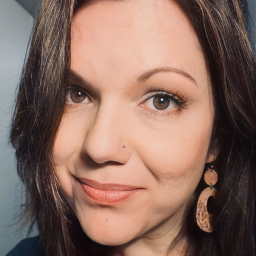 The image shows Shana looking at the camera with a slight smile. She has dark brown shoulder-length hair, brown eyes, and a nose piercing on her left nostril. She is wearing a black sweater and brown wooden dangly earrings.