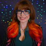 A person with long red, orange and pink hair with large blue glasses who is smiling wearing a black and green collared jacket. The background is grey with rainbow lights in the distance.