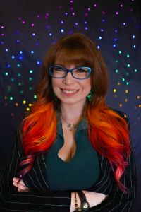A person with long red, orange and pink hair with large blue glasses who is smiling wearing a black and green collared jacket. The background is grey with rainbow lights in the distance.