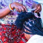 Edna Shaibu está sentada en el suelo e inclina su cabeza sobre los muslos de la peluquera para que le trencen el cabello, en un salón ubicado en la calle Buswelu, en la región de Mwanza, Tanzania. La peluquera colocó una toalla blanca sobre los hombros de Edna para evitar que trozos de cabello caigan sobre su vestido. Su cabeza, con el cabello parcialmente trenzado, está expuesta a la cámara. La foto fue tomada por George Binagi el 4 de noviembre de 2024.