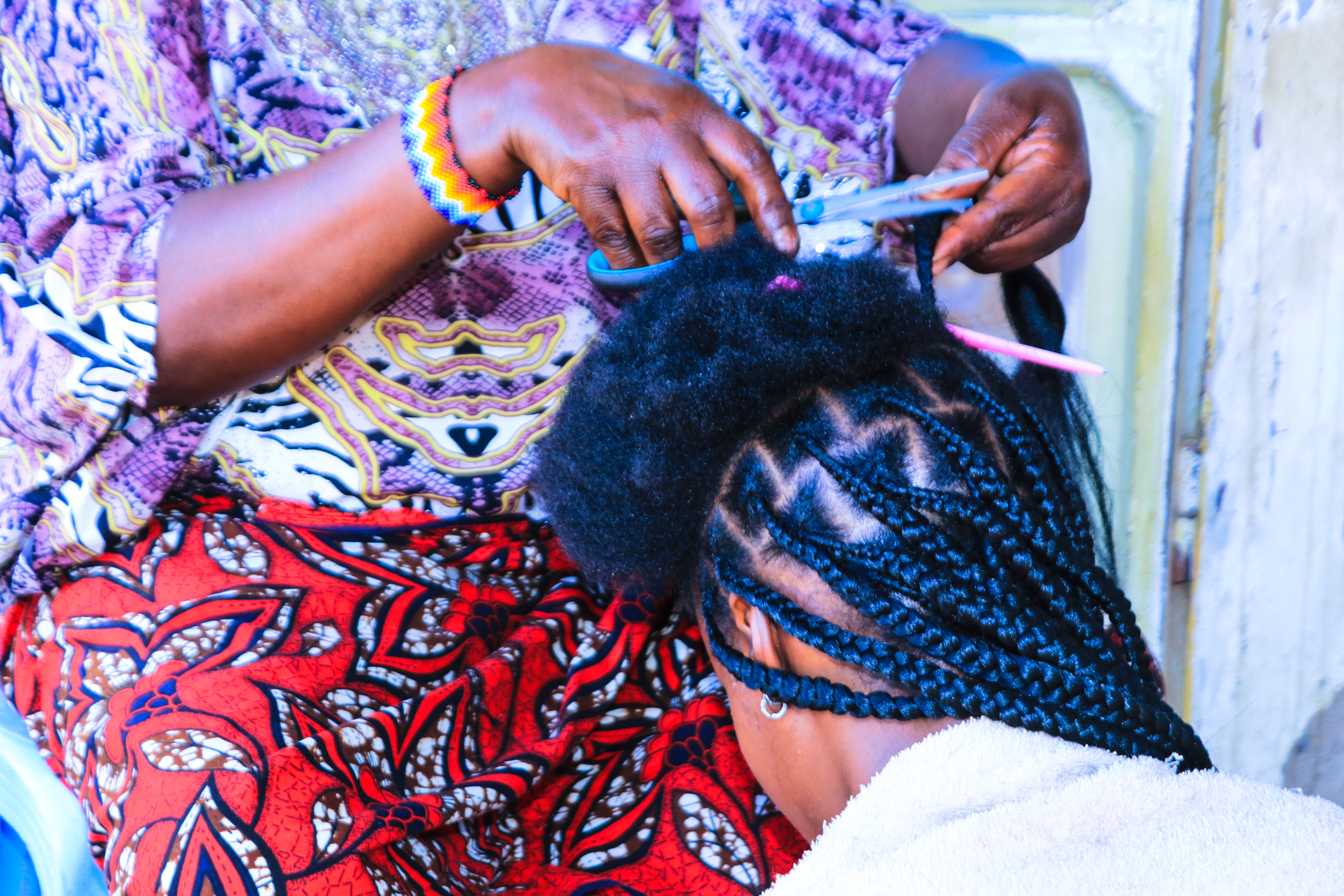 Edna Shaibu está sentada en el suelo e inclina su cabeza sobre los muslos de la peluquera para que le trencen el cabello, en un salón ubicado en la calle Buswelu, en la región de Mwanza, Tanzania. La peluquera colocó una toalla blanca sobre los hombros de Edna para evitar que trozos de cabello caigan sobre su vestido. Su cabeza, con el cabello parcialmente trenzado, está expuesta a la cámara. La foto fue tomada por George Binagi el 4 de noviembre de 2024.
