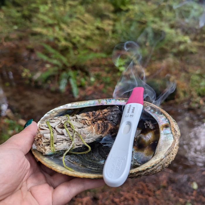 Una mano sostiene una concha de abulón durante una ceremonia de sahumerio. Dentro de la concha hay pasto dulce, salvia y una prueba de embarazo. La foto fue tomada por Seren Friskie el 16 de noviembre de 2024.