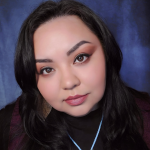 Headshot of Johnnie Jae, a Choctaw and Otoe-Missouria with black hair parted to the side, dark brown eyes with a slight smirk as she stares at the camer with her head slightly tilted. She is wearing a Black tee with a dark violet red sweater vest and beaded cat bolo tie.