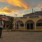 En la imagen aparece la cineasta Ángeles Cruz en un atardecer de colores cafés y amarillos, de pie frente a la Agencia Municipal de su pueblo Villa Guadalupe Victoria. Se está haciendo una trenza mientras mira hacia la cámara. Crédito de la foto: Ange Cayuman, 5 de enero de 2022