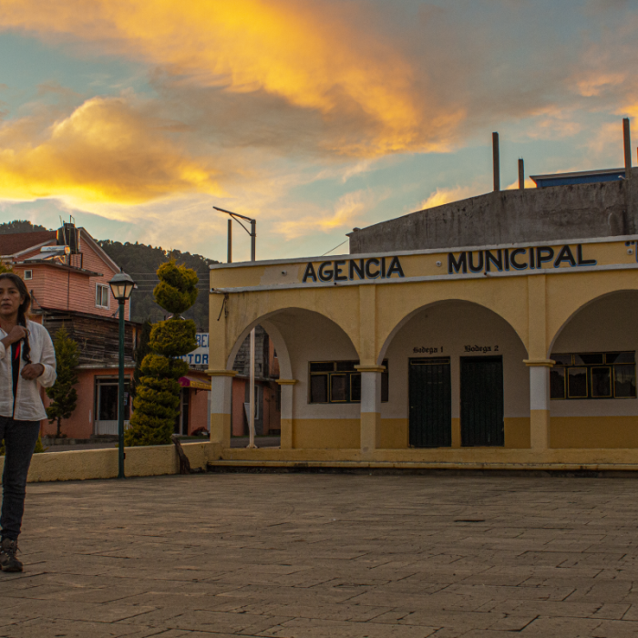 En la imagen aparece la cineasta Ángeles Cruz en un atardecer de colores cafés y amarillos, de pie frente a la Agencia Municipal de su pueblo Villa Guadalupe Victoria. Se está haciendo una trenza mientras mira hacia la cámara. Crédito de la foto: Ange Cayuman, 5 de enero de 2022