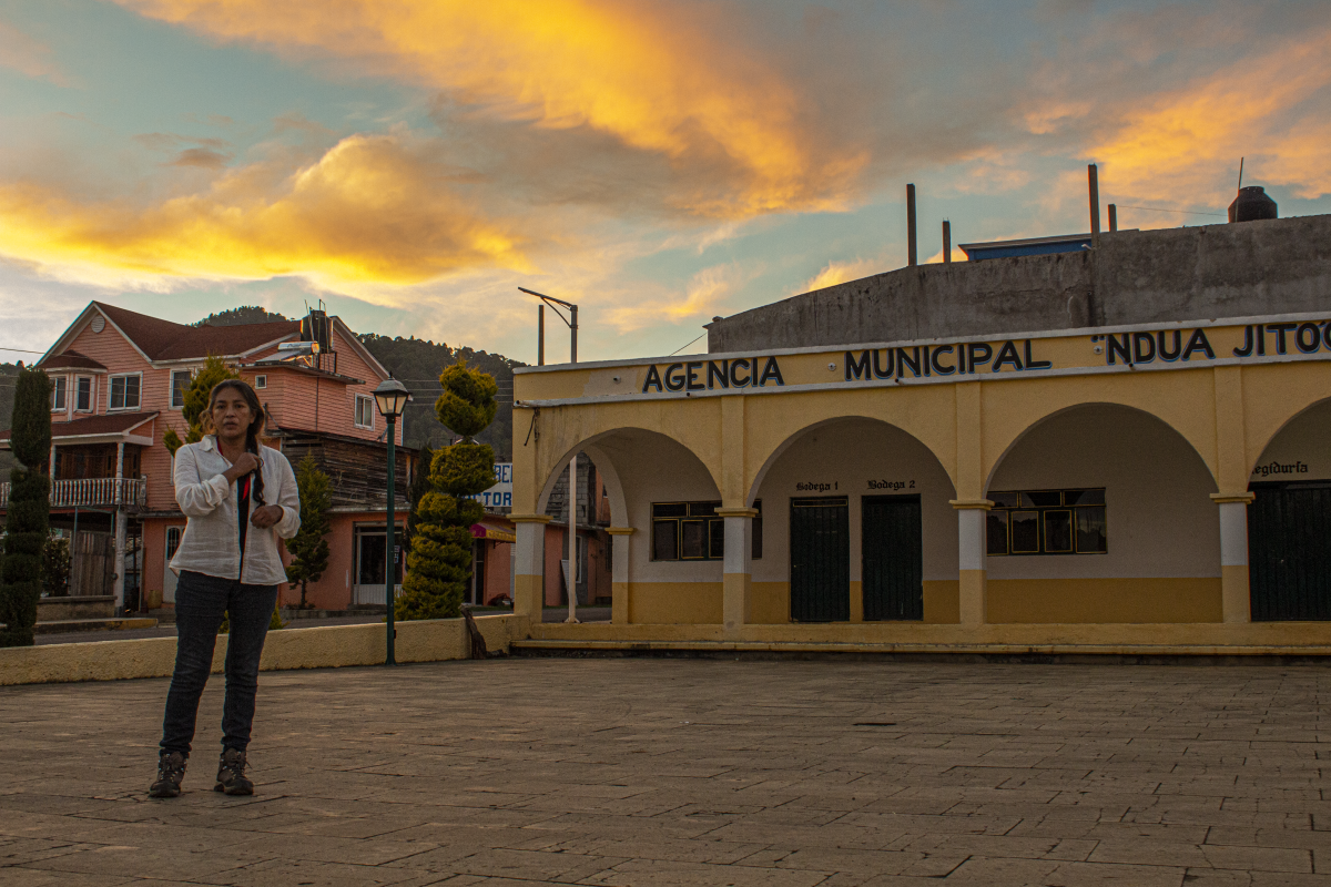 En la imagen aparece la cineasta Ángeles Cruz en un atardecer de colores cafés y amarillos, de pie frente a la Agencia Municipal de su pueblo Villa Guadalupe Victoria. Se está haciendo una trenza mientras mira hacia la cámara. Crédito de la foto: Ange Cayuman, 5 de enero de 2022