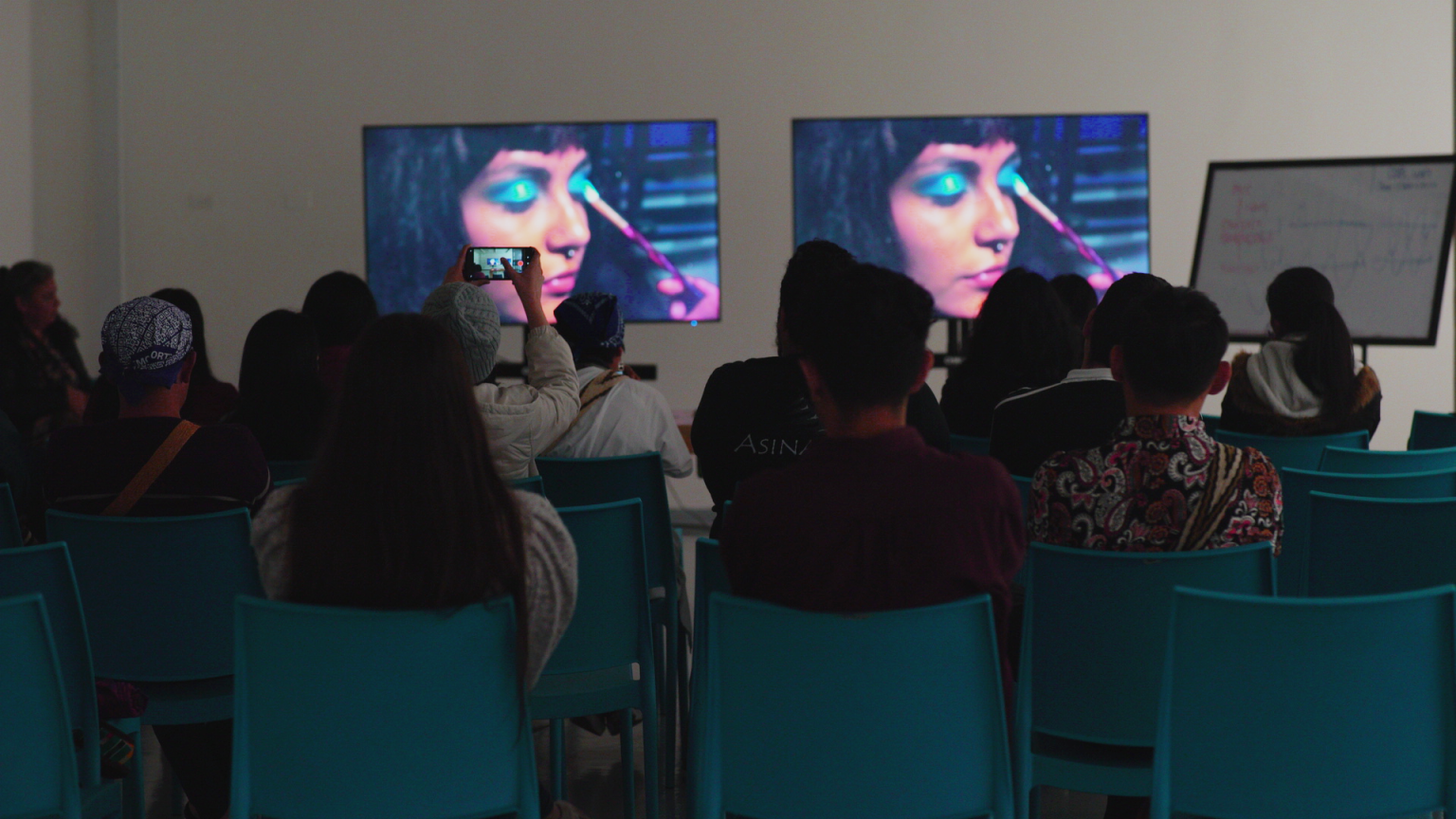 La foto muestra personas sentadas en sillas azules observando una imagen proyectada en una pared, un fotograma del video “Eymi Inchiw”, del artista Pablo Lincura, en el que aparece una joven pintándose los labios. La muestra es parte de la muestra de cine de Diversidades Ancestrales en Daupará. Crédito de la foto: Ange Cayuman, 18 de octubre de 2022