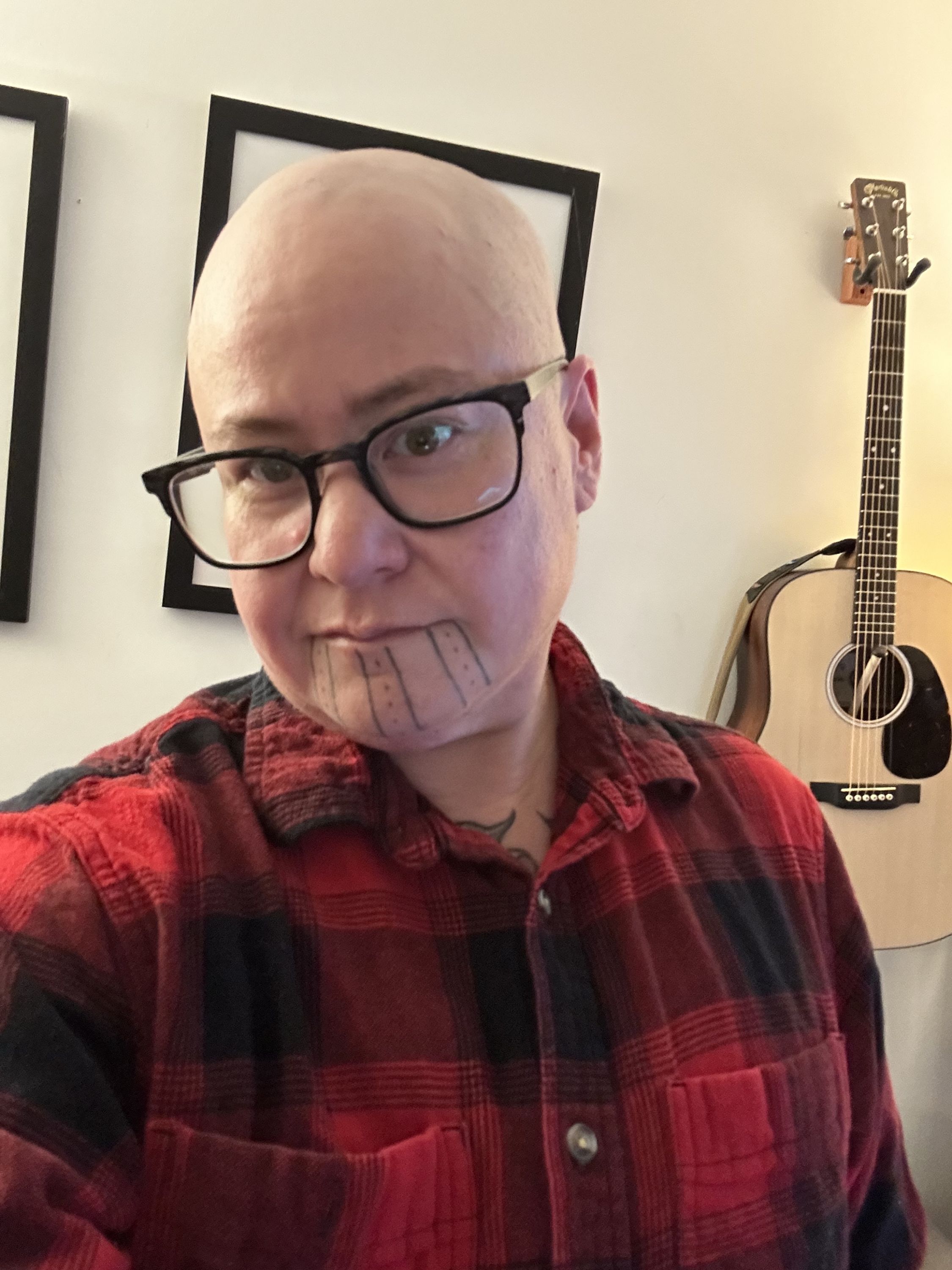 Un hombre trans indígena de piel clara, con la cabeza rapada, está de pie en su sala usando una camisa de cuadros rojos y negros. Hay una guitarra en la pared y una lámpara encendida detrás de él. Crédito de la foto: Theo Jean Cuthand, 2025