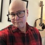 Un hombre trans indígena de piel clara, con la cabeza rapada, está de pie en su sala usando una camisa de cuadros rojos y negros. Hay una guitarra en la pared y una lámpara encendida detrás de él. Crédito de la foto: Theo Jean Cuthand, 2025