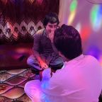 Two people sit on a carpeted floor in a cozy, warmly lit room with multicolored lights reflecting on the walls. One of the persons, named Annie, is wearing beige grey shalwar kameez, and smiles gently while her partner, dressed in white, holds her hand affectionately.