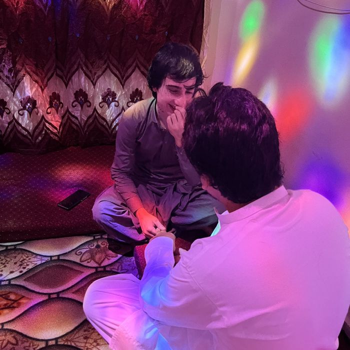 Dos personas están sentadas sobre una alfombra, en una habitación acogedora y cálidamente iluminada, con luces multicolores reflejándose en las paredes. Una de ellas, llamada Annie, viste un shalwar kameez beige y gris. Sonríe con dulzura mientras su pareja, vestida de blanco, le toma la mano con cariño.