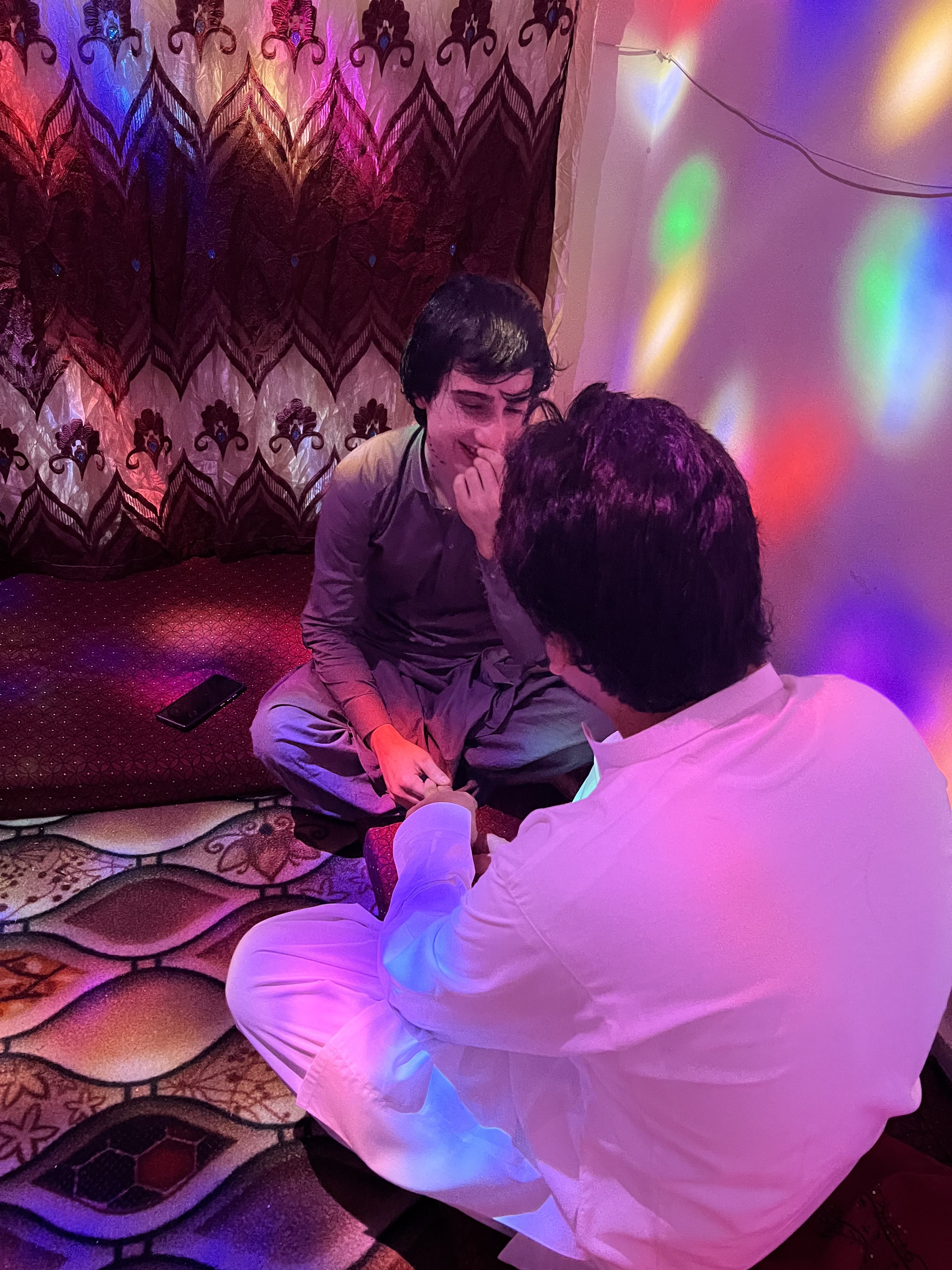 Dos personas están sentadas sobre una alfombra, en una habitación acogedora y cálidamente iluminada, con luces multicolores reflejándose en las paredes. Una de ellas, llamada Annie, viste un shalwar kameez beige y gris. Sonríe con dulzura mientras su pareja, vestida de blanco, le toma la mano con cariño.