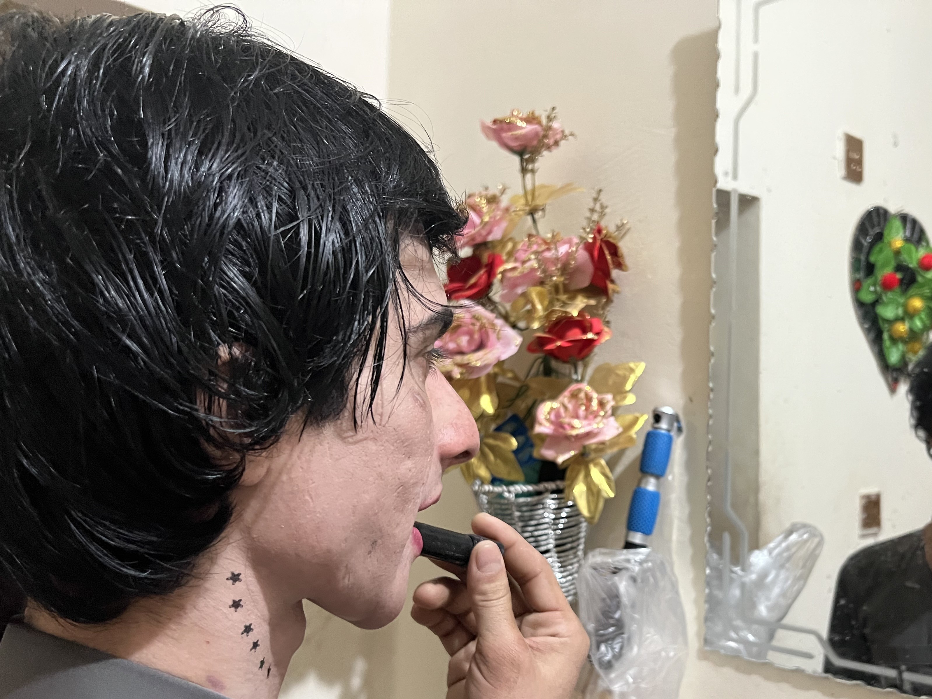 Annie, con el cabello negro y mojado, está de pie frente a un espejo, pintándose los labios. Tiene una pequeña hilera de tatuajes de estrellas en el cuello y viste un traje gris beige. En el reflejo del espejo, su expresión es concentrada. El estante debajo del espejo contiene artículos de maquillaje y junto a ellos se ve un arreglo floral decorativo.