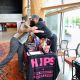 Two individuals share a warm embrace. One person stands behind a table covered with a black cloth printed with the acronym “HIPS” in large pink and white letters, along with the phrases “Honoring Individual Power & Strength” and “HARM REDUCTION SAVES LIVES.” They wear a black cap and black face mask. The other person leans forward into the hug, dressed in a striped shirt and gray pants. The table is arranged with informational pamphlets, candy, and promotional materials. The venue features large windows, wooden columns, seating areas, and potted plants, creating a bright and welcoming atmosphere.