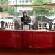 A jewelry display booth is set up indoors. The booth is covered with a red tablecloth and showcases a variety of jewelry pieces, including necklaces, bracelets, and earrings, all arranged neatly on display stands and trays. A woman with medium-size dark hair wearing a face mask and glasses stands behind the booth. Large glass windows in the background reveal an urban street scene with parked cars and buildings, adding depth and context to the indoor setting.