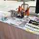 A display table is set up. The table is covered with a white cloth and neatly arranged with various items. These include multiple copies of a publication titled “THE MAGAZINE” with the subtitle “CRUSHING COLONIALISM,” white and black cloths, which may be t-shirts or flags printed with bold black or white and yellow text reading “CRUSHING COLONIALISM,” and a circular stickers that says “DECOLONIZED PRIDE”. Small rainbow flags are scattered across the table. Also present are brochures and flyers with Indigenous themes, a bottle of hand sanitizer, individually wrapped candies, and a few cylindrical containers with printed designs.