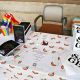 A display table is set up. The table is covered with a white cloth featuring a rainbow pattern and is neatly arranged with various materials. These include a container filled with small rainbow pride flags, several black booklets with colorful illustrations and a white building on the cover, and a stack of white booklets titled “SAGE”. A printed flyer sits in the center of the table. A chair is positioned behind the table.